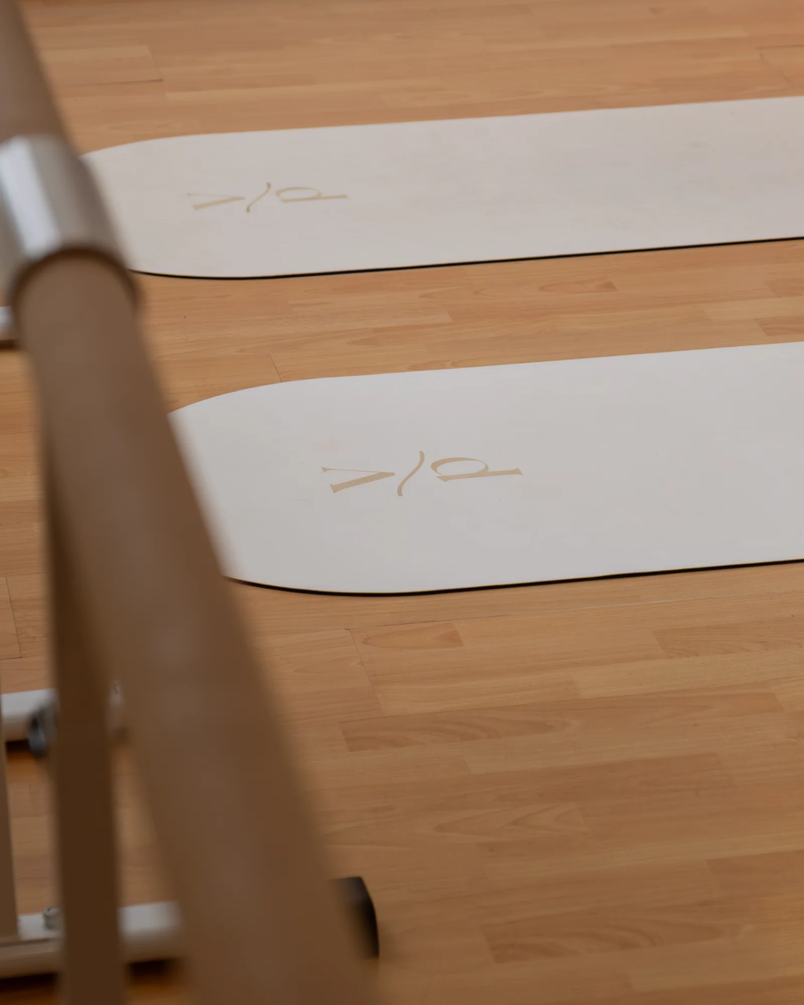 Two cream exercise mats stamped with the studio monogram, laid out on pale timber flooring beyond the blurred rails of a barre.