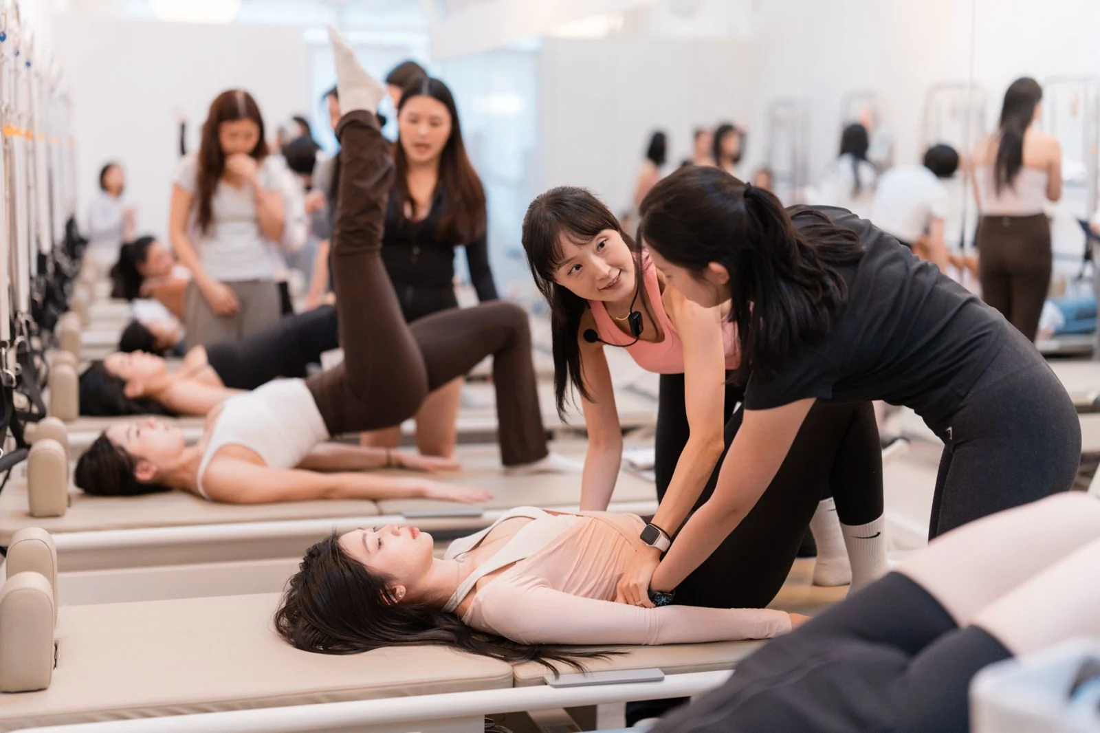 An instructor in a pink top, headset mic on, coaches a trainee in black as she presses a hand to the hip of a student lying on a reformer, a full training room working behind them.