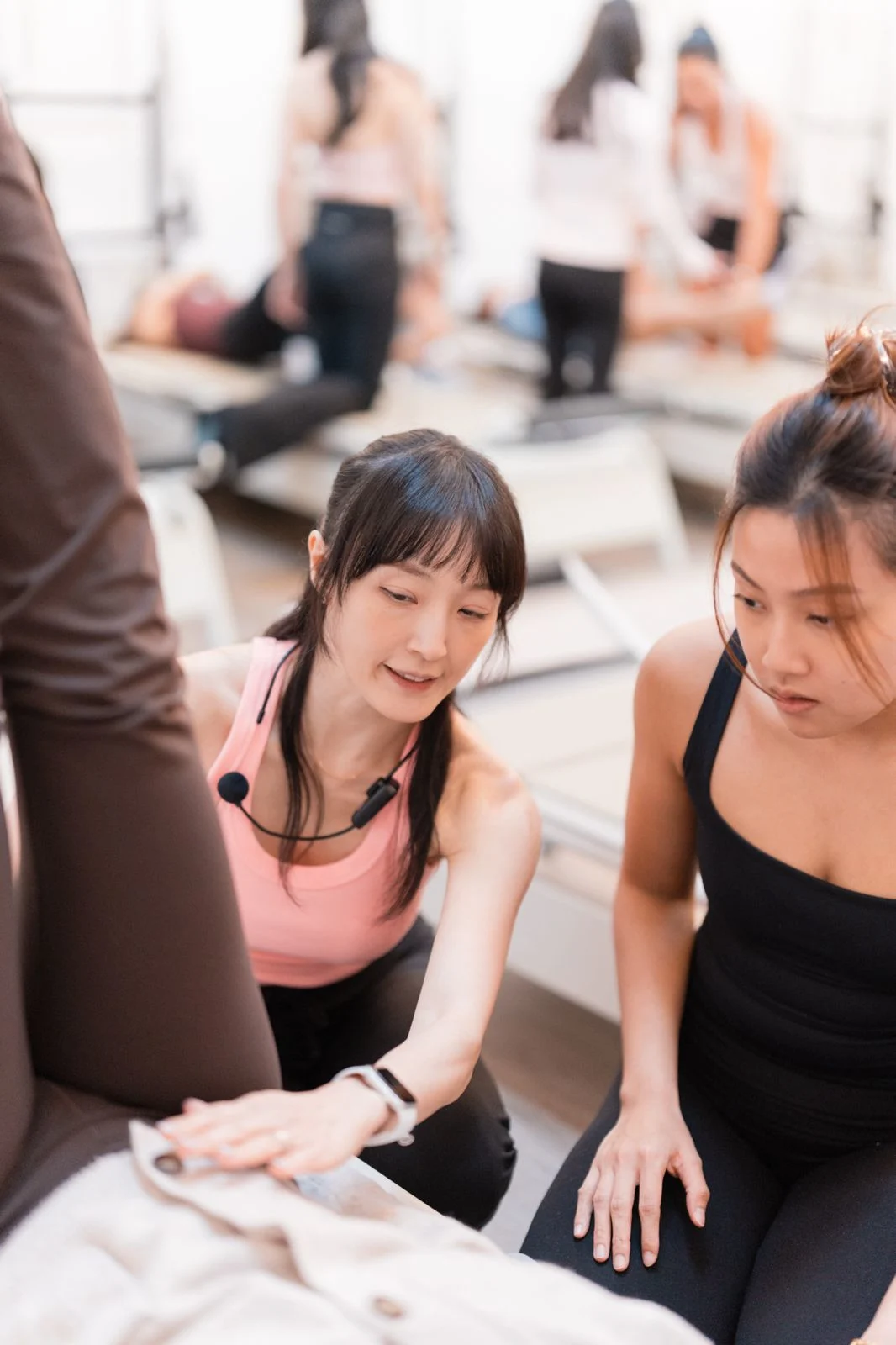 Close in on the instructor in pink, headset mic on, resting a hand on the reformer beside a student's raised leg as a trainee in black crouches beside her and watches.