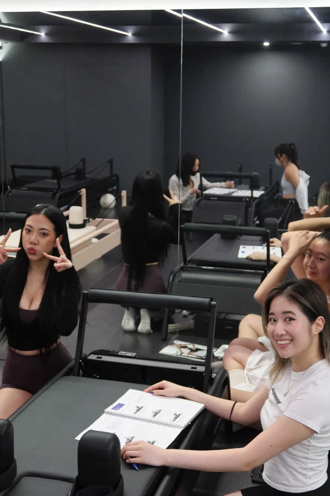 Trainees at black reformers in a dark-walled studio, course manuals open in front of them, two turning to the camera with a smile and a peace sign.