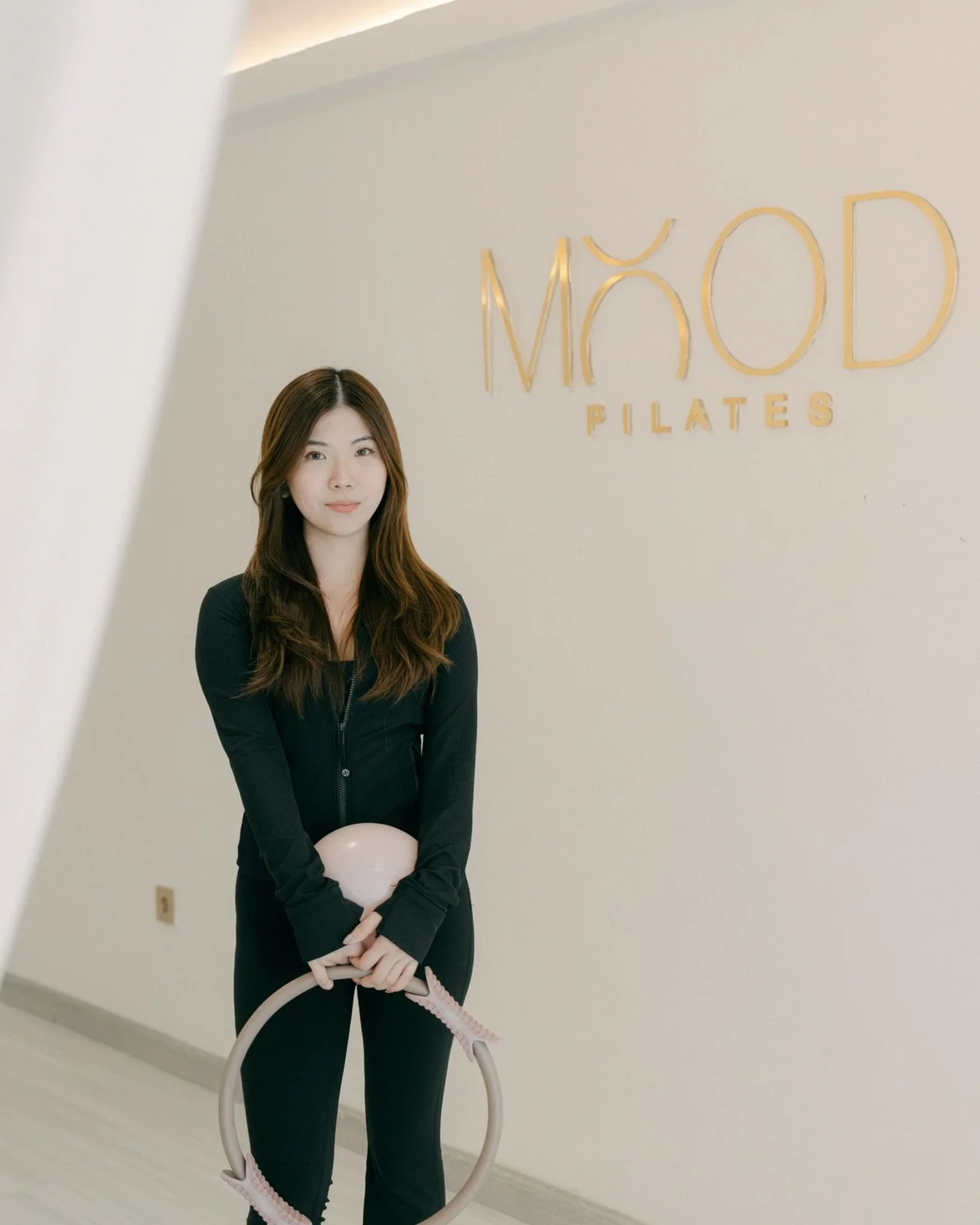 Celine holding a Pilates ring and small ball in her studio, the gold Mood Pilates lettering on the wall beside her.