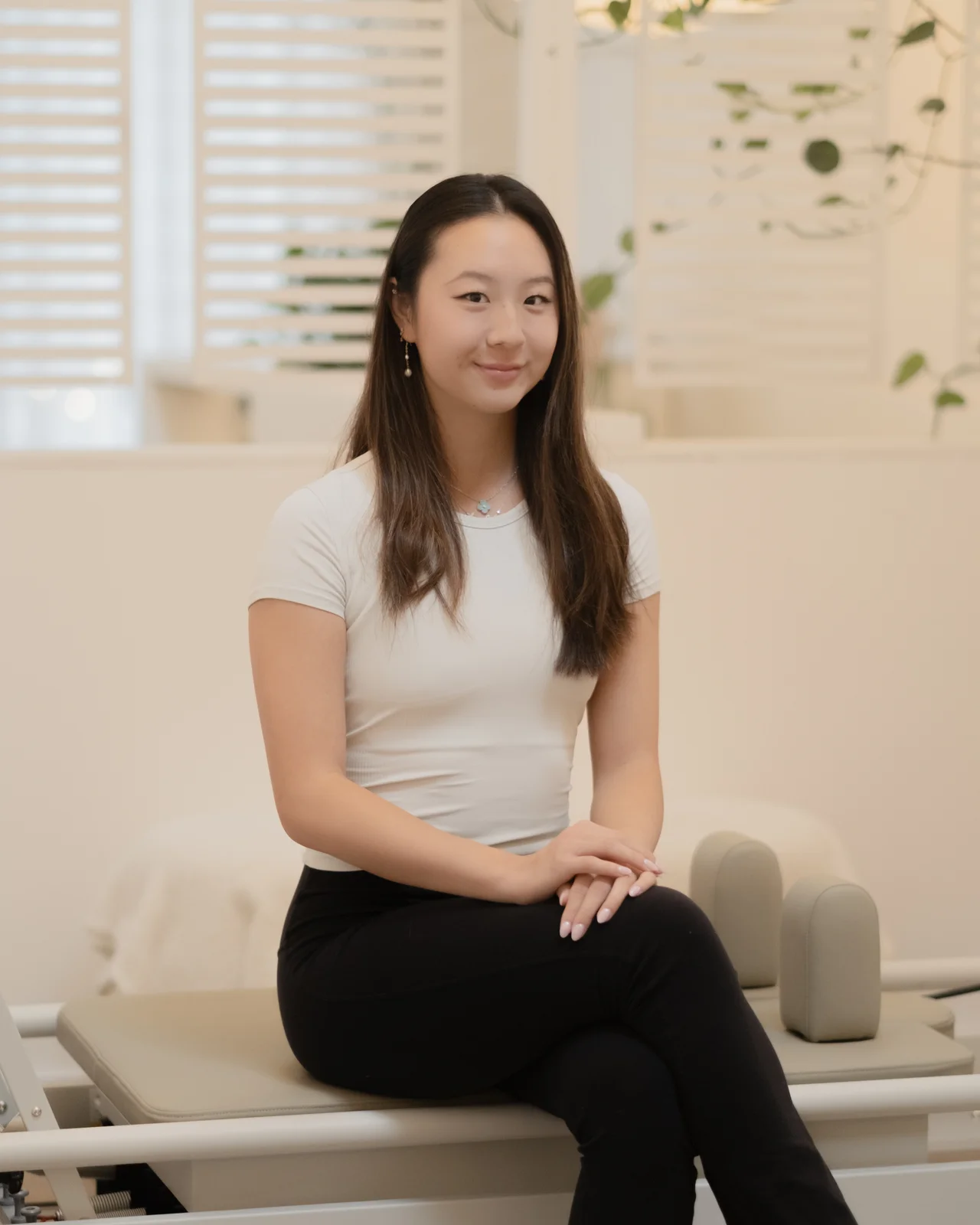 Vivian seated on a reformer bed in a cream studio, hands resting on her knee, smiling at the camera.