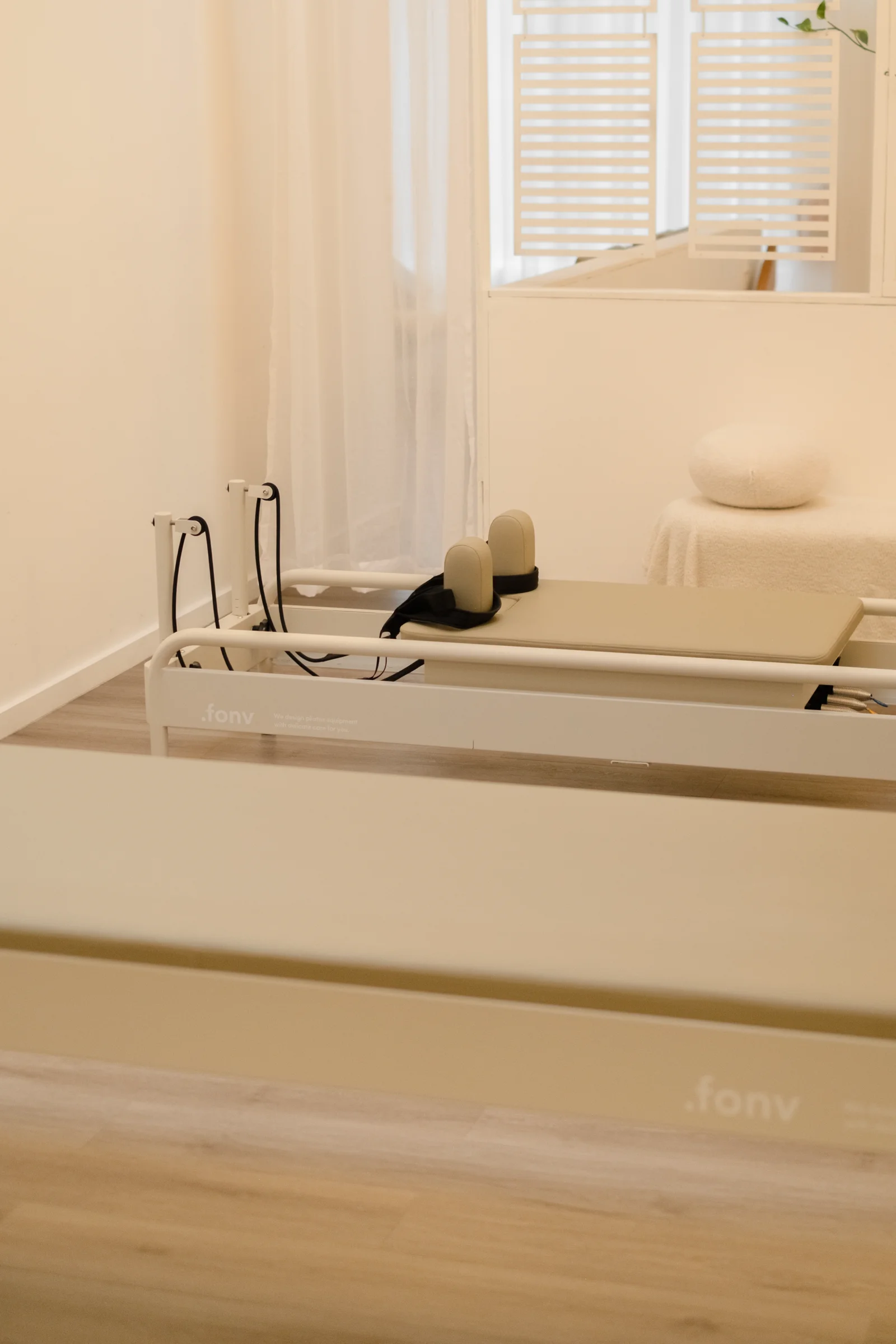 Empty cream reformer beds stamped with the fonv wordmark, lined up on pale timber toward a sheer-curtained window and a massage table with a round bolster.