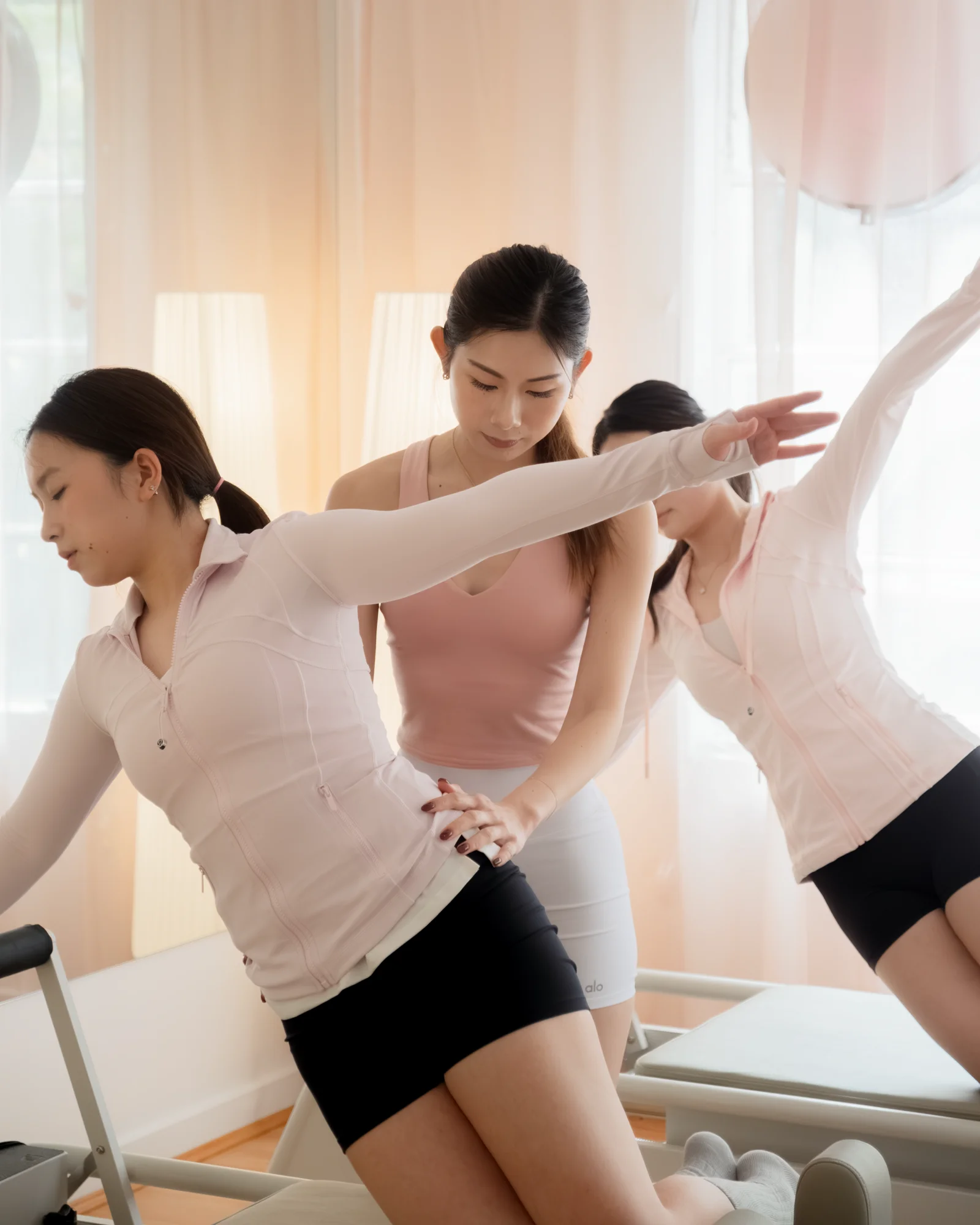 An instructor in blush guides a student's raised arm and hip through a kneeling side bend at the reformer, a second student mirroring the reach behind sheer curtains.