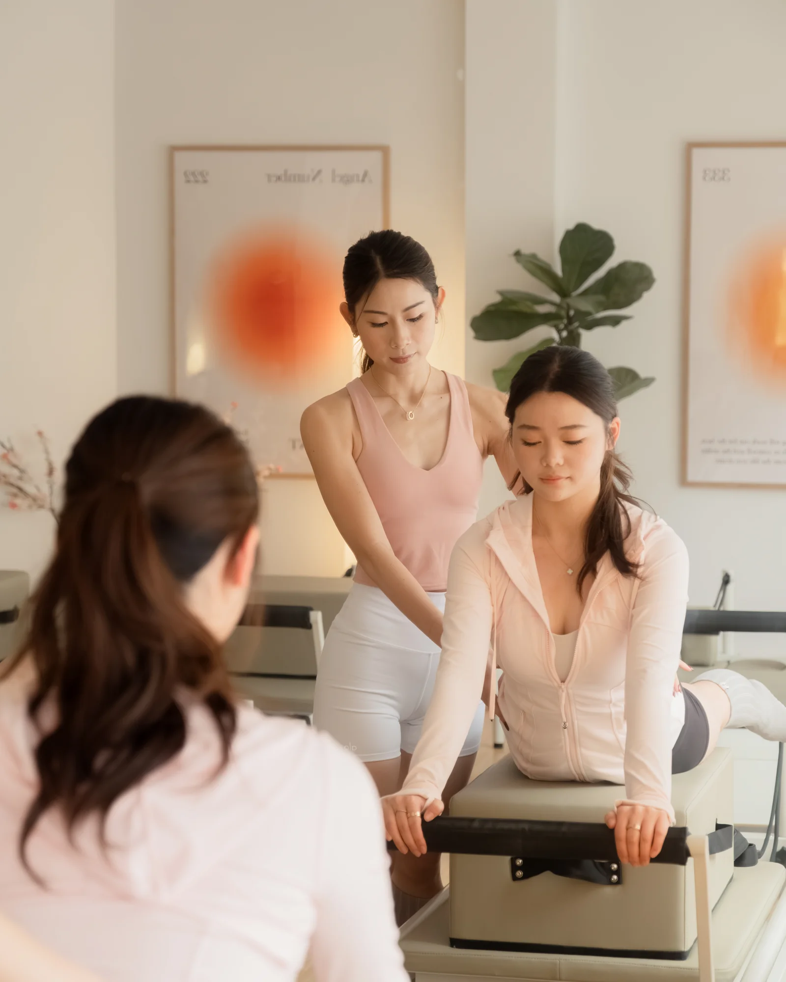 An instructor guides a client through a supported stretch over a reformer box, hands at her shoulders, while another client looks on from the foreground.