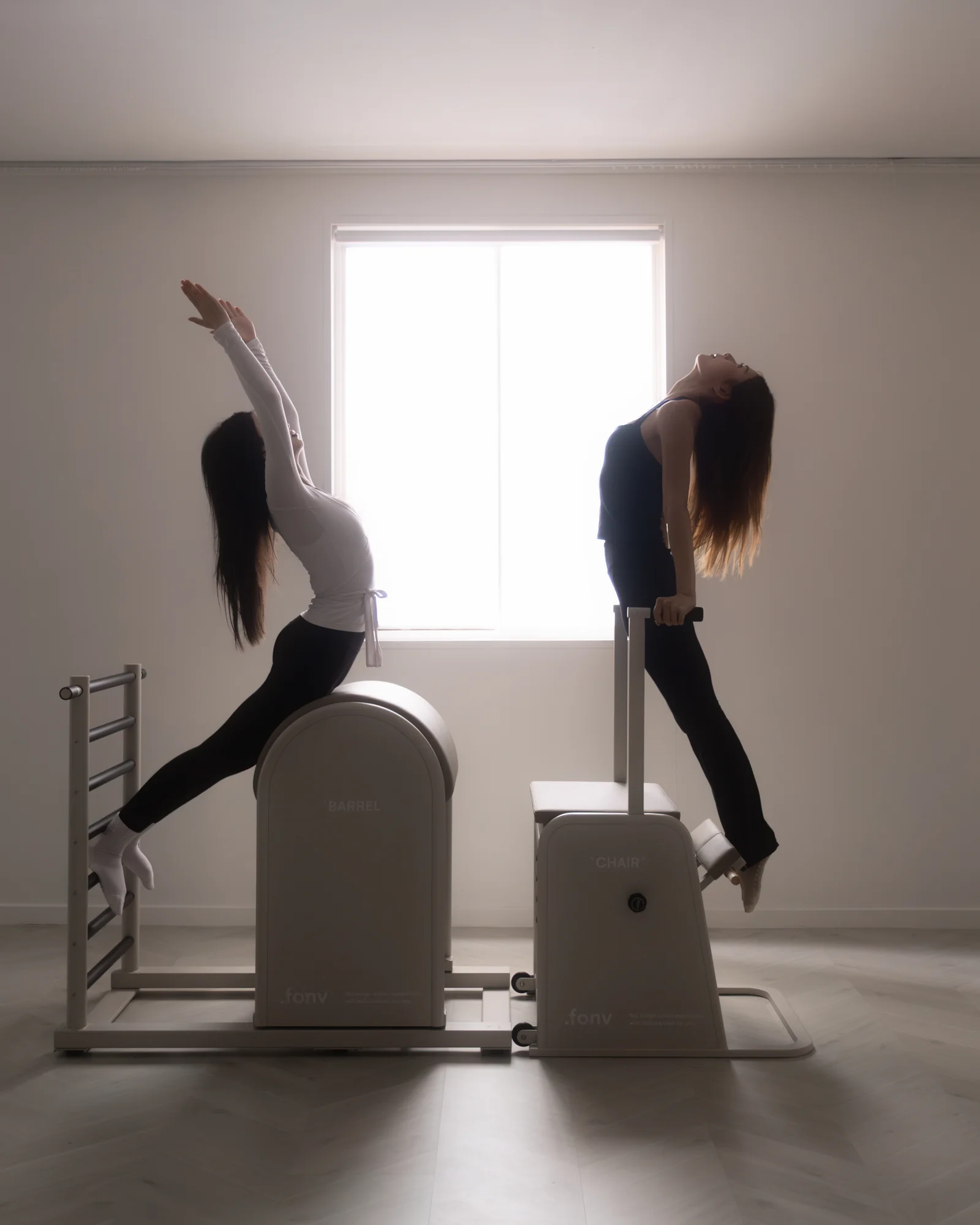 Two women silhouetted against a bright studio window — one reaching into a lunge stretch over a ladder barrel, the other arching back atop a wunda chair.
