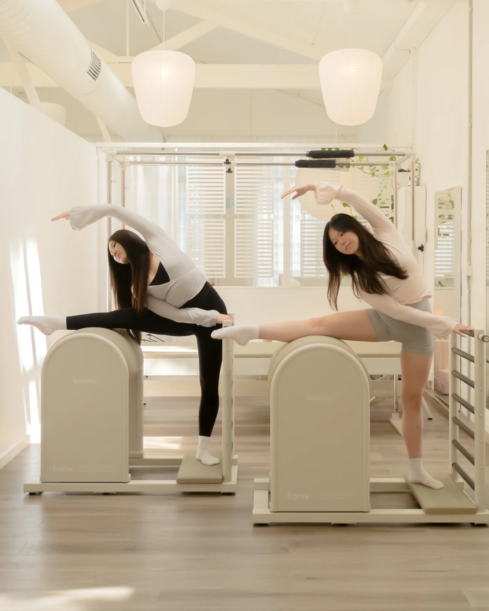 Two women mirror a side bend over ladder barrels, one leg extended along the barrel top, beneath white paper lanterns.