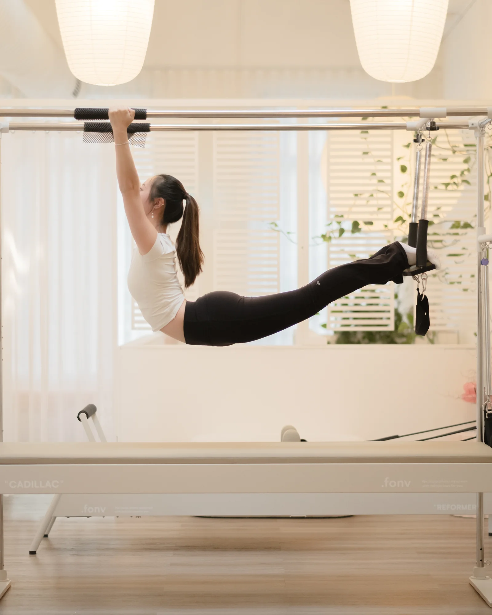 A client hangs from a Cadillac top bar, body stretched into one long horizontal line, feet hooked into a strap behind her.